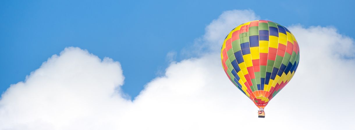 Solitary hot air balloon floating among the white, puffy clouds against a bright blue sky.