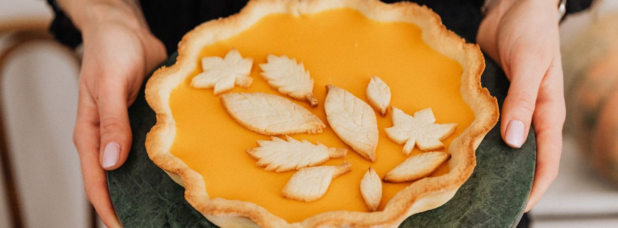 Woman's hands shown holding a pumpkin pie topped with pastry leaves on a serving dish.